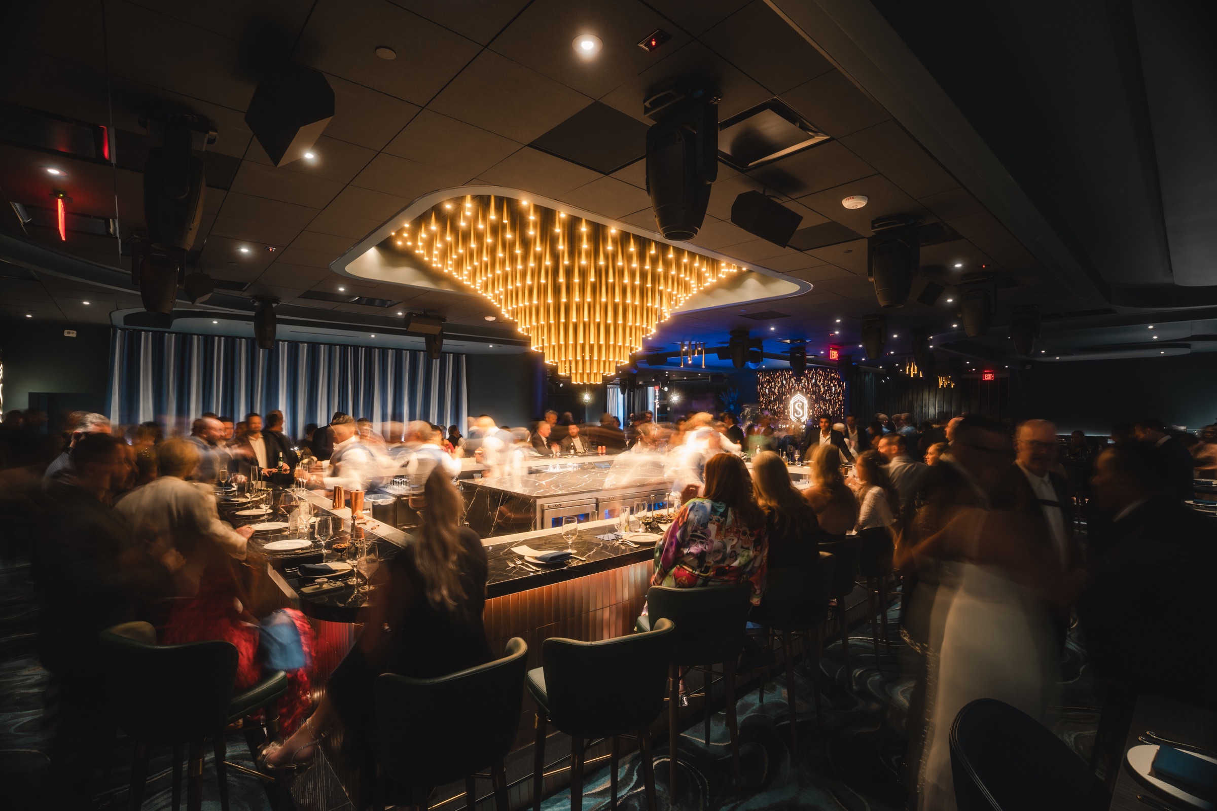 Full restaurant room in motion beneath a sculptural gold light installation