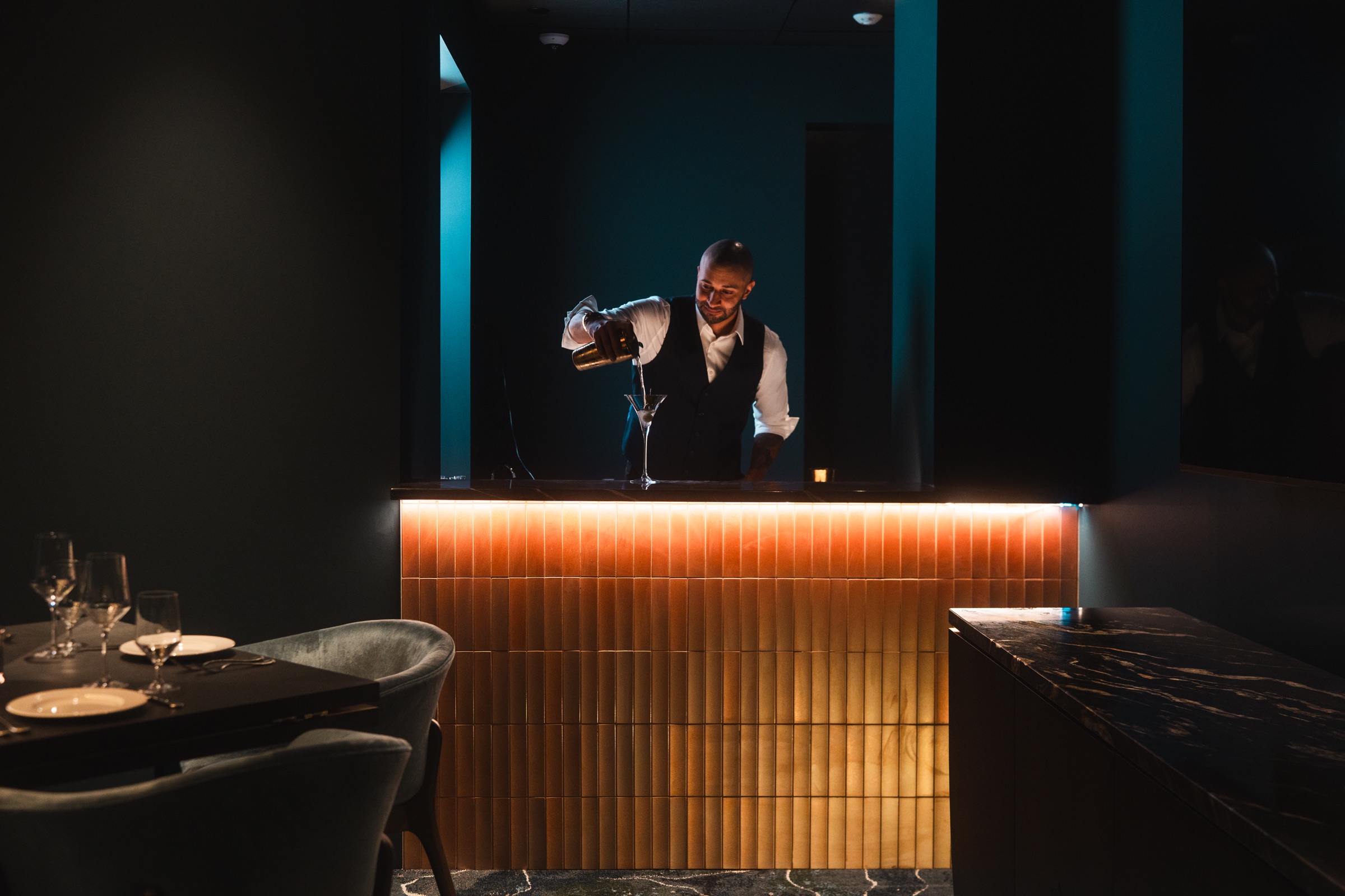 Bartender pouring a cocktail behind a warmly lit bar in a dark, elegant dining room