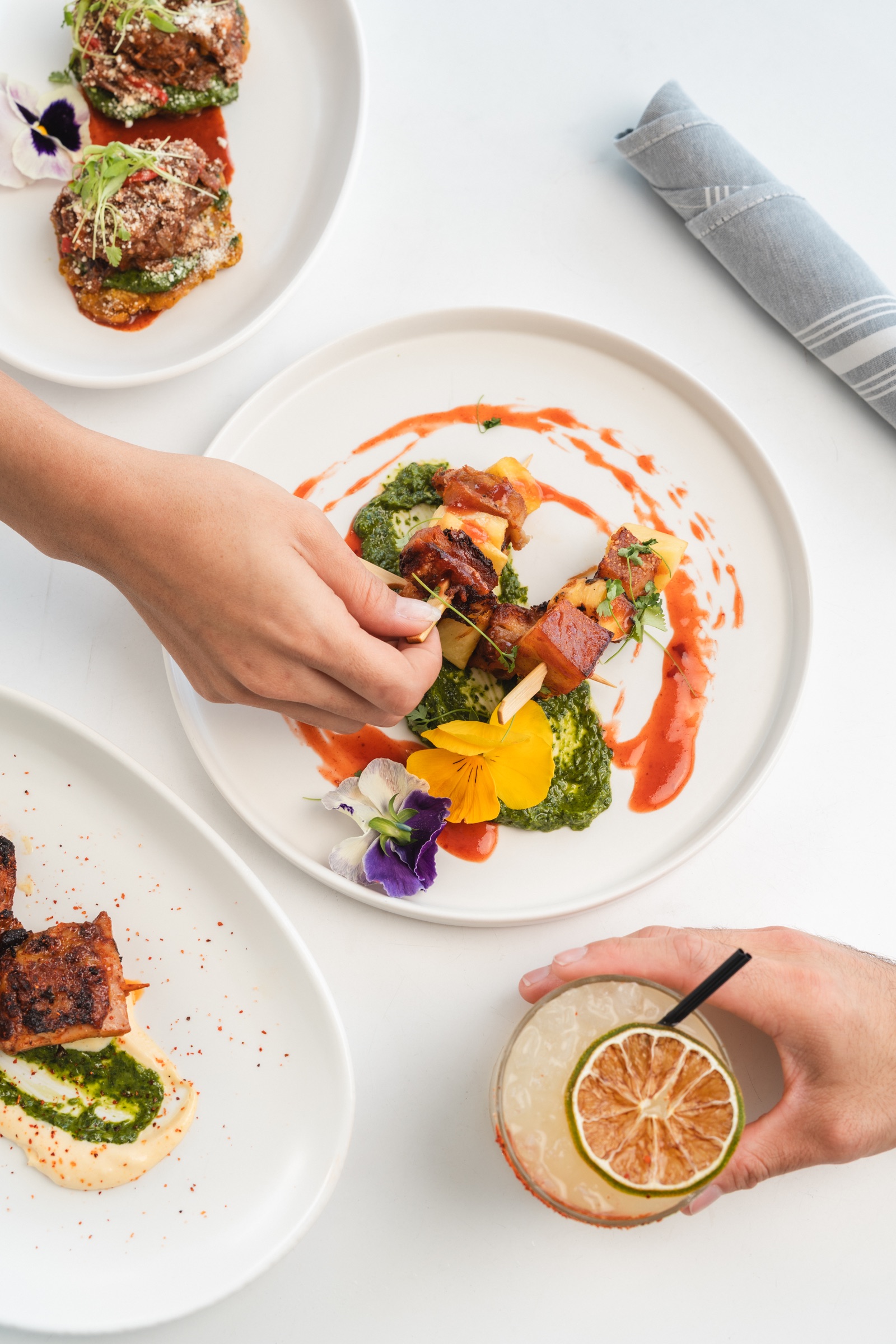 Bright overhead table scene with plated dishes, hands, and a cocktail